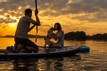 Happy couple paddle boarding at lake during sunset together with pug dog. Concept of active family...