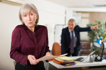 Portrait of frustrated elderly female office employee standing with paper in hands on background of disgruntled manager reprimanding her - obrazy, fototapety, plakaty