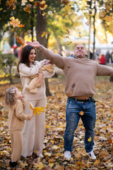 Young happy family with two children in an autumn park.  Mom holds her youngest daughter in her arms, and dad throws leaves up.  Everyone is dressed in a beige color scheme