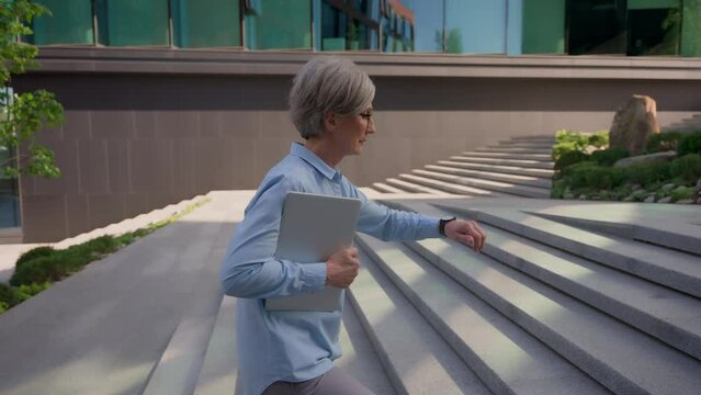 Caucasian businesswoman adult business employer mature senior woman hold folder walk up stairs office building in city middle-aged female looking at wrist watch time late hurry rush running lateness