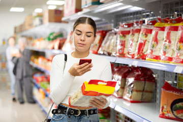 Girl buyer examines packaging of product and scan QR code on label. Young woman in supermarket view and buys daily groceries, pasta Chinese noodles