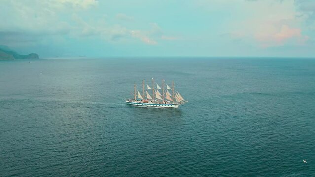 A large multi-masted sailing ship cruises the serene blue sea, with a clear sky above and calm waters around. - Powered by Adobe
