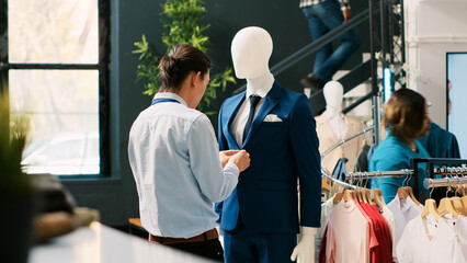 Stylish man checking mannequin with formal suit, arranging fashionable clothes before store opening. Asian employee working with trendy merchandise and accessories in modern boutique. Fashion concept