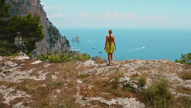 Girl hiking on Capri Island and gazing from a cliff edge on sea below. Woman enjoying view of vastness of the seascape, with the endless ocean meeting the sky. Italy.