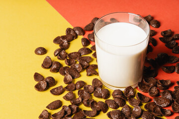 Chocolate corn flakes scattered on a yellow-red background, fresh milk poured into a glass