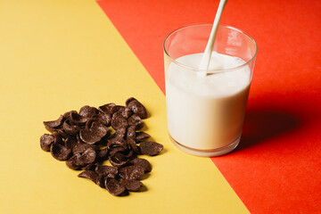 Chocolate corn flakes scattered on a yellow-red background, next to the glass fresh milk poured...