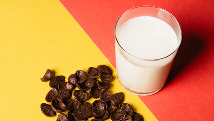 Chocolate corn flakes scattered on a yellow-red background, fresh milk poured into a glass