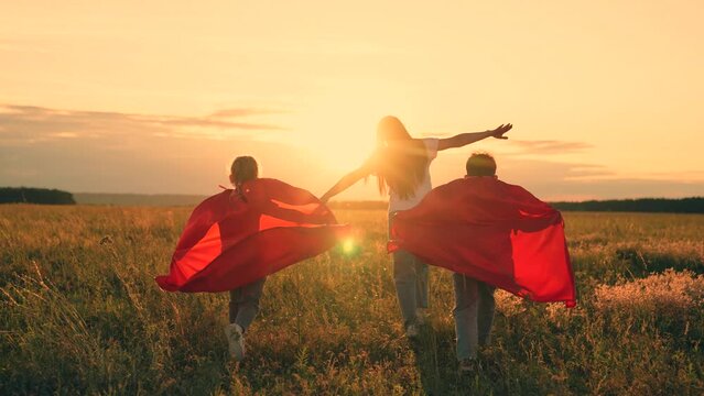 Mother daughter, son play superheroes in park. Mom child run in red raincoats, have fun outdoor. Family Carnival in sun, Halloween, sky. Mom, Super kids dream of becoming superhero, flying in red cape