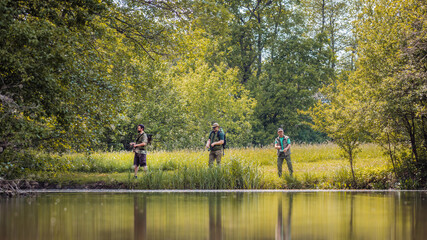 Three sports fishermen standing on the river bank, under a beautiful tree, swinging their fishing rods trying to catch fish on a sunny summer day.