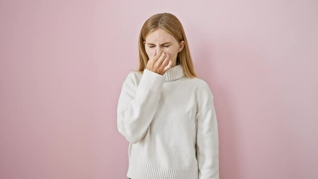 Disgusting stench overpowers beautiful blonde girl, unhappy expression as woman holds breath, pinching nose amid intolerable smell. standing isolated on pink background with sweater.