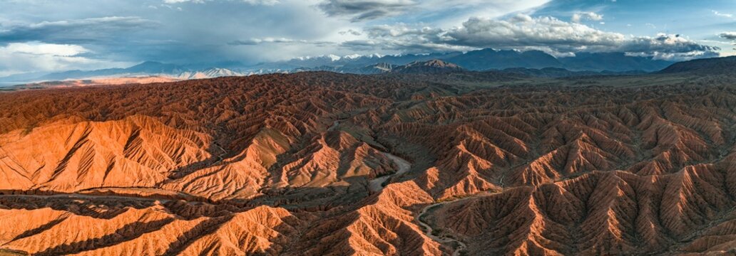 River bed runs through a landscape of eroded hills, badlands at sunset, mountain peaks of the Tian Shan Mountains in the background, aerial view, Canyon of the Forgotten Rivers, Issyk Kul