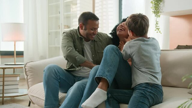 Happy family African American dad and little boy Caucasian son tickling mom at home playing funny game laughing relax on couch together adorable child kid having fun with father mother love children