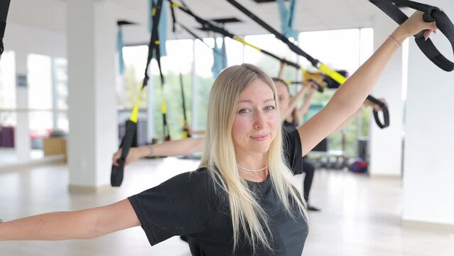 Women in gym using TRX equipment for joint, arm, leg, and chest exercises.