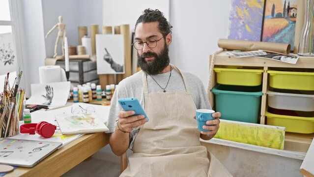 A cheerful bearded man enjoys coffee and smartphone in a bright art studio