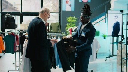African american store assistant helping regular client with a suit in shopping center, senior customer looking to buy formal clothes. Elderly man shopping for new collection at the mall. Camera B.