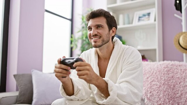 A relaxed man in a white robe plays a video game in a cozy, modern living room, exemplifying leisure and technology at home.
