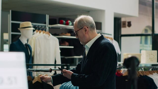 Trendy senior man shopping for new fashion collection at retail store, visiting modern clothing shop to check formal or casual items. Rich customer uses phone to look for products by code. Camera A.