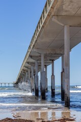 Portion of Concrete Pier Crossing Frame From Right to Left, with Waves Crashing into Pylons and Distant Surfers, Shot from Underneath, San Diego, California, USA, vertical