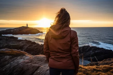Tuinposter Woman in a navy windbreaker enjoying a solitary moment on a rocky shoreline as the sun sets behind a distant lighthouse © aicandy