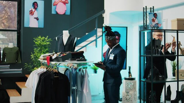 African american worker checking inventory for new clothing line placed on hangers for sale, looking at merchandise on tablet and in store. Retail assistant verifying discount list at mall. Camera B.