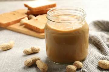 Tasty peanut butter in glass jar and peanuts on gray table, closeup