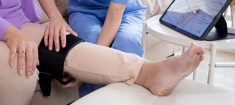 Caregiver or nurse helping check tendon and arthritis of knee or leg for diagnostic and rehabilitation while explaining senior woman with tablet living room at home, caretaker or physiotherapist.