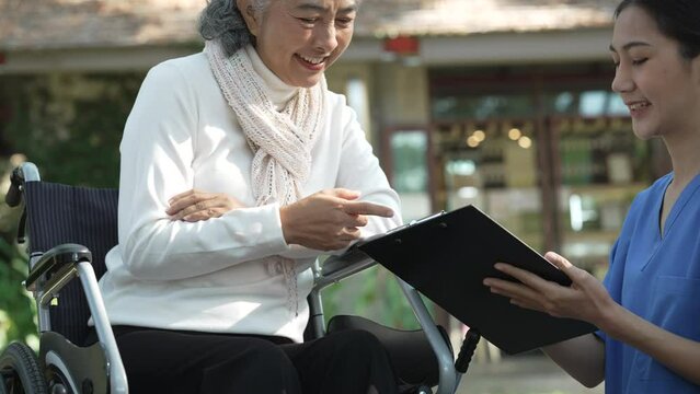 Compassionate Asian woman provides care to  elderly person in wheelchair outdoors. Engaging in physical therapy, happiness, encouraging positive environment for mature individuals with grey hair.