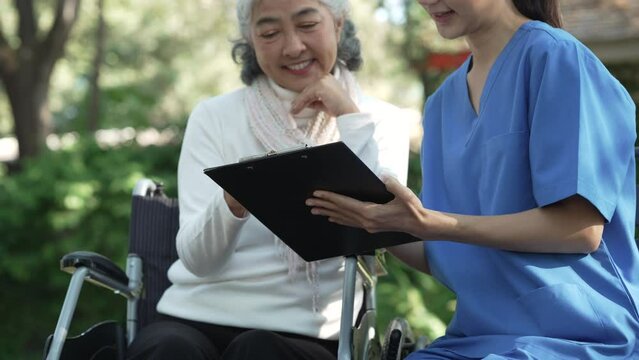 Compassionate Asian woman provides care to  elderly person in wheelchair outdoors. Engaging in physical therapy, happiness, encouraging positive environment for mature individuals with grey hair.