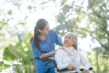 Compassionate Asian woman provides care to  elderly person in wheelchair outdoors. Engaging in...
