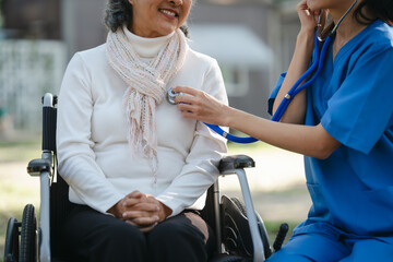 Compassionate Asian woman provides care to  elderly person in wheelchair outdoors. Engaging in...