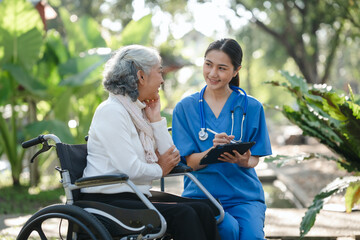 Naklejka premium Compassionate Asian woman provides care to elderly person in wheelchair outdoors. Engaging in physical therapy, happiness, encouraging positive environment for mature individuals with grey hair.