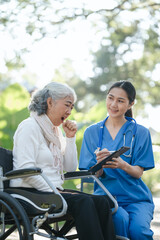 Compassionate Asian woman provides care to  elderly person in wheelchair outdoors. Engaging in physical therapy, happiness, encouraging positive environment for mature individuals with grey hair.