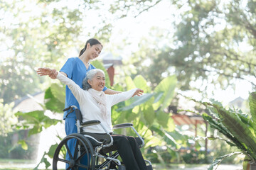 Compassionate Asian woman provides care to  elderly person in wheelchair outdoors. Engaging in physical therapy, happiness, encouraging positive environment for mature individuals with grey hair.