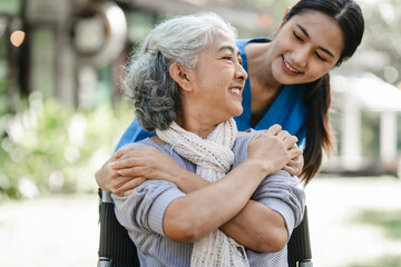 Compassionate Asian woman provides care to  elderly person in wheelchair outdoors. Engaging in physical therapy, happiness, encouraging positive environment for mature individuals with grey hair.