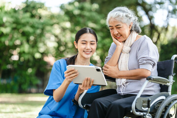 Compassionate Asian woman provides care to  elderly person in wheelchair outdoors. Engaging in...