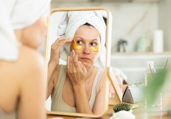 Relaxed woman in her middle age using under eye patches for skincare sitting at the mirror in home...