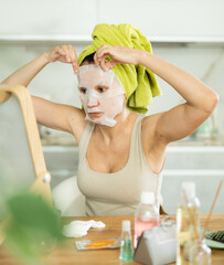 Obraz na płótnie Canvas Careful middle-aged lady doing face care with sheet mask while seated at a table in her house