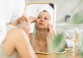 Calm middle-aged woman examining her facial skin looking in the mirror put on the table in her...