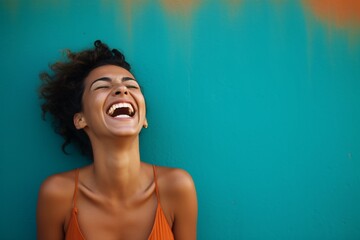 Black beautiful model teenage woman with curly hair dressed in orange tak top laughing and smiling in front of a teal wall, funny, happiness, joy, enjoy life	