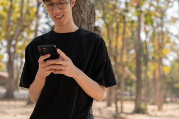 A positive young Asian man using his smartphone in a park, chatting with his friends.