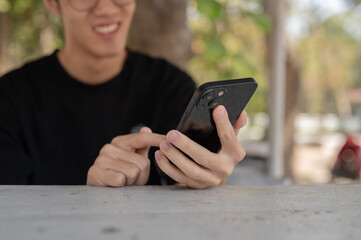 A happy Asian man chatting with his friends on his smartphone while sitting at a table outdoor.