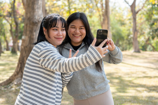 Two young happy Asian female best friends taking selfies with a smartphone together in a park.