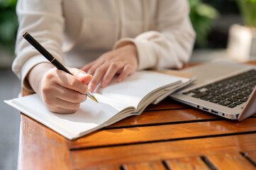 Fototapeta premium A woman in a white hoodie working remotely at an outdoor cafe, listing her ideas in a book