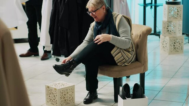 Retail worker presenting various pairs of shoes for senior client, assisting woman to find the perfect footwear in fashion showroom. Elderly customer trying on footgear before buying. Camera A.