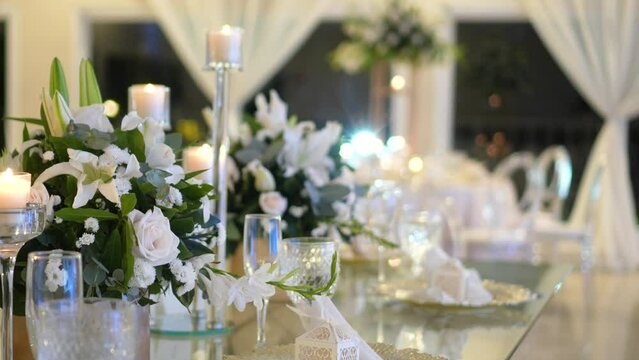 Glass table decorated with centerpieces with natural foliage and white roses, candlesticks and glassware for wedding reception.