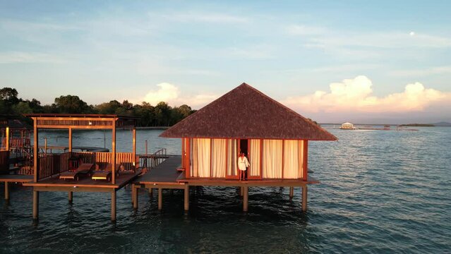 Over Water Bungalow Resort at Leebong Island, Belitung Indonesia - Woman Traveler Opens Villa Doors, Walks Out and Sits on Deck Edge To Watch Sunset - Aerial Pull Back