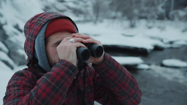 Male hiker in outerwear looking through binoculars from the bank of mountain river during winter travel in nature