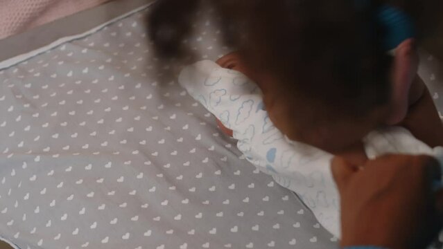 Over shoulder of unrecognizable African American woman putting white onesie with light blue clouds print on her newborn son on changing table at home