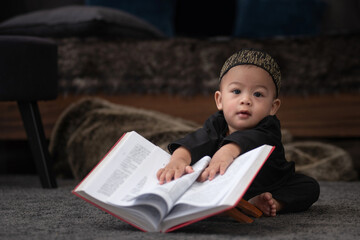 Cute Muslim child boy in traditional clothes is interested in the Qur'an, sitting on the floor pretending to read the Quran at home, looking at camera