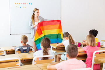Schoolchildren sitting in classroom and listening to female teacher. She holding rainbow flag in hands and talking about minorities.